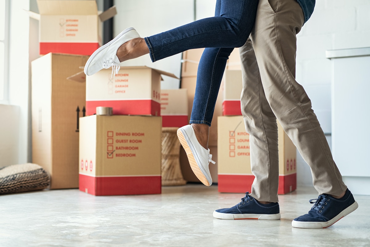 Man lifting his girlfriend among cardboard boxes as they start living together in new house. Closeup of excited husband embracing wife celebrating relocation. Loving man standing near boxes lifting woman up in the air excited about moving in.